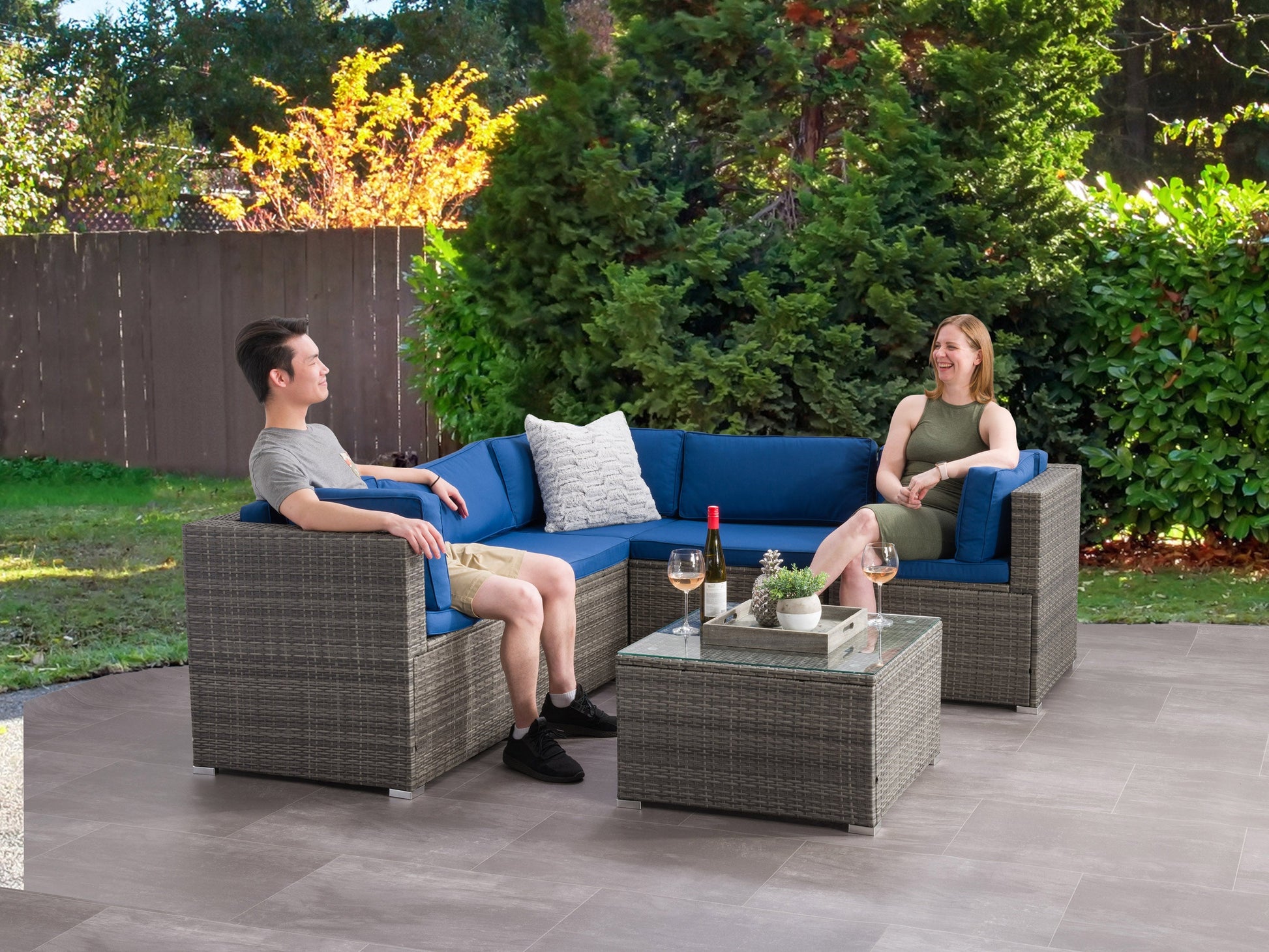 Gray patio sectional with plush cushions, woven rattan frame, and glass-top coffee table on a wooden deck, set against a lush garden backdrop. Perfect for outdoor lounging and entertaining.