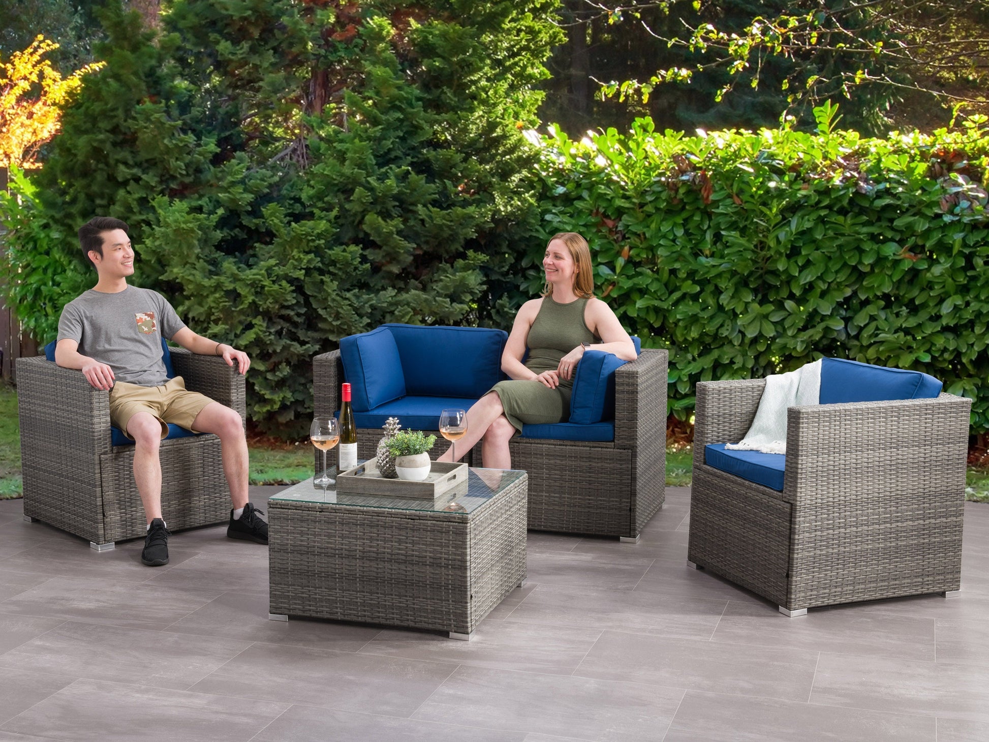 Modern patio conversation set with gray wicker sofa, matching armchairs, and a glass-top coffee table, featuring plush white cushions, set on a wooden deck with green plants in the background.